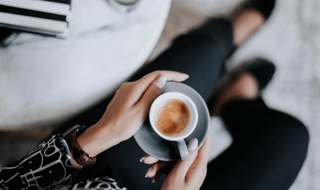 business woman sits next to a marble table with books and holds a saucer with a cup of espresso